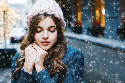 Frau mit warmer Mütze, die in winterlicher Umgebung steht und die Hände vor der Brust hält, während Schnee fällt, symbolisiert die Notwendigkeit von Hautpflege im Winter.