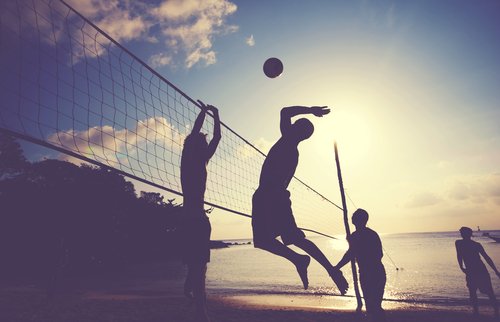 Silhouetten von Beachvolleyballspielern, die vor dem Hintergrund eines Sonnenuntergangs springen und einen Ball schmettern, was Outdoor-Sport und Sonneneinstrahlung betont.