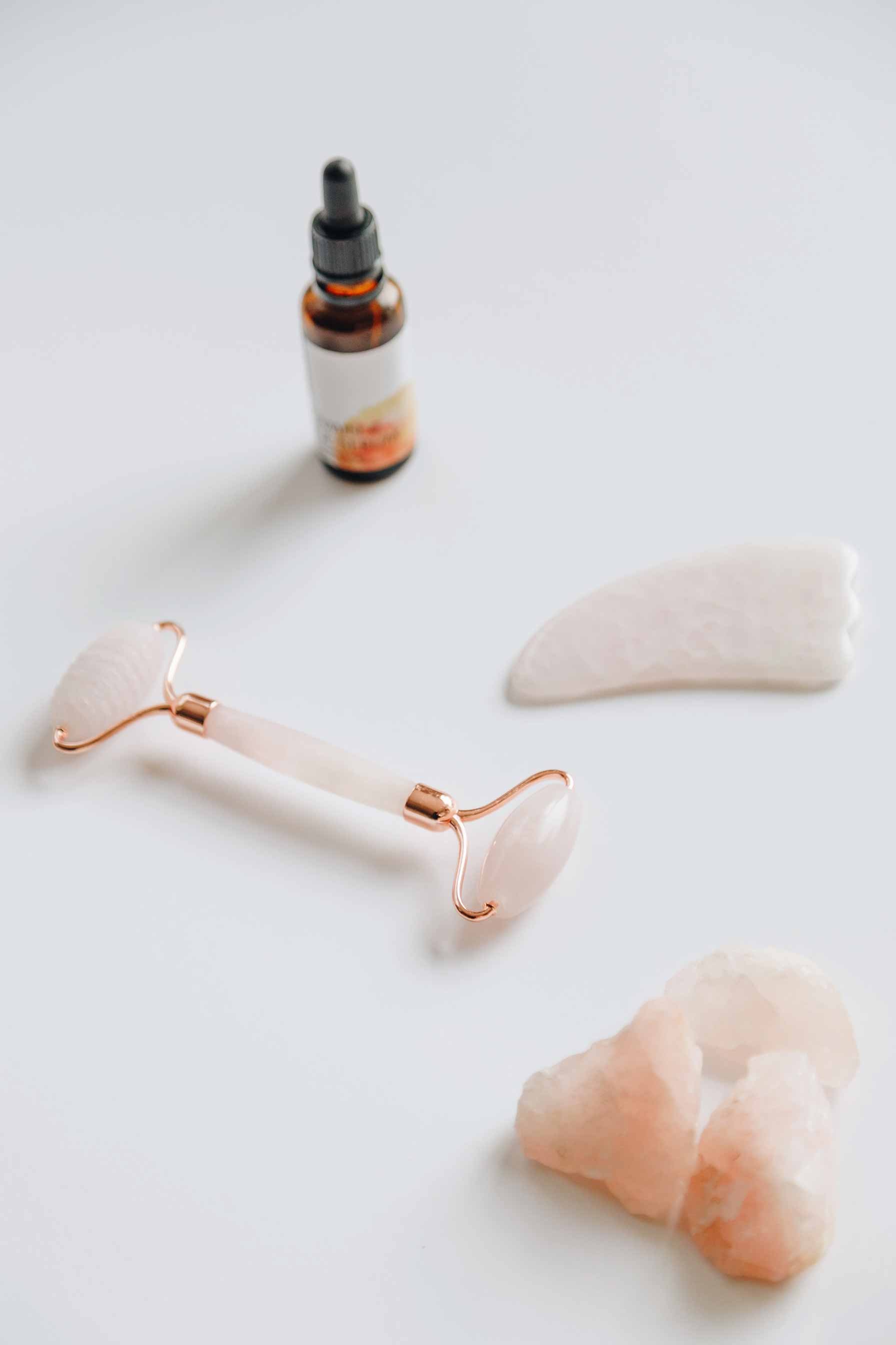 Rosenquarz Gesichtsroller, Gua Sha Stein und Hautpflegeöl auf weißem Hintergrund, symbolisierend die Hautpflege-Trends 2020 und natürliche Beauty-Rituale.
