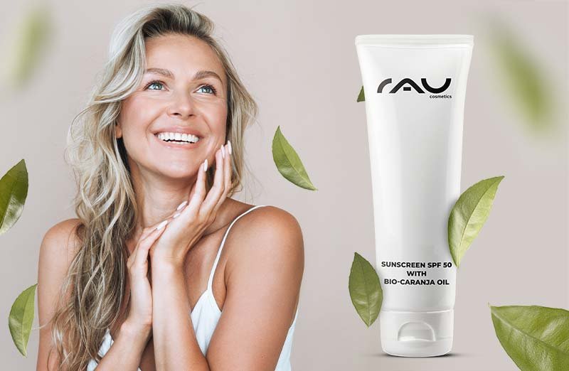 Frau mit freudigem Gesicht und Sonnenschutzmittel SPF 50 mit Bio-Karanja-Öl von RAU Cosmetics vor grünen Blättern.