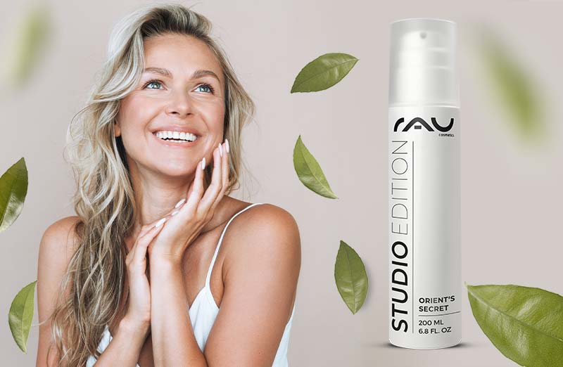 Lächelnde Frau mit blonden Wellenhaaren hält "Orient's Secret" Gesichtscreme von RAU Cosmetics vor grünem Hintergrund.