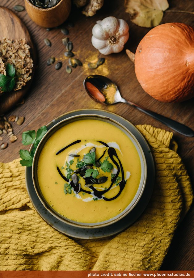 Kürbissuppe in einer Schüssel, garniert mit Kräutern, Kürbiskernen und einem Spritzer Kürbiskernöl, umgeben von frischen Kürbissen, Knoblauch und Gewürzen auf einem rustikalen Holzuntergrund.