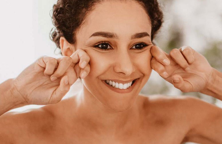 Frau mit fröhlichem Gesichtsausdruck, die ihre Wangen mit den Händen zieht, zur Veranschaulichung von manueller Lymphdrainage zur Reduzierung von Wassereinlagerungen im Gesicht.