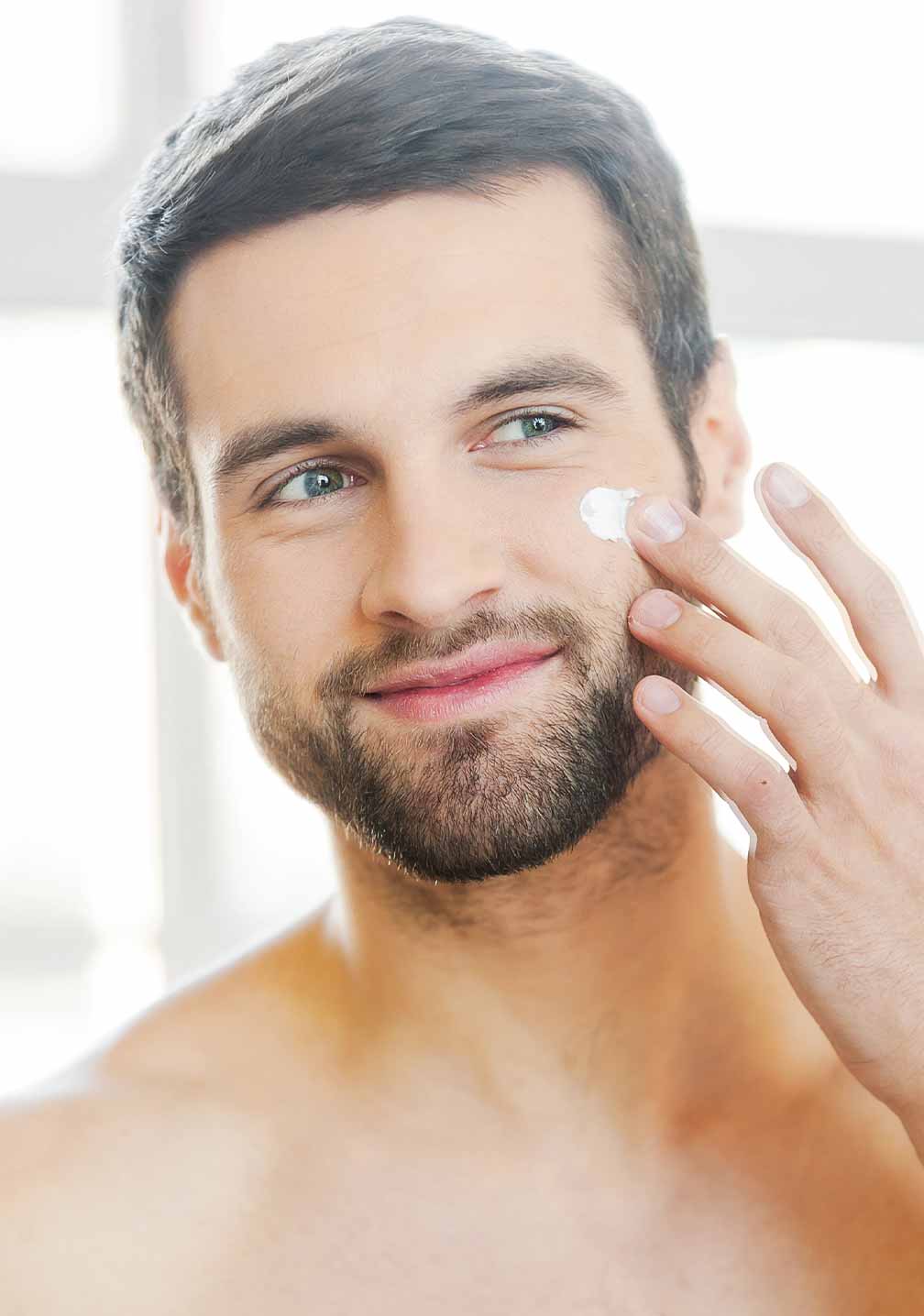 Junger Mann mit gepflegtem Bart, der RAU Cosmetics XXXY Lotion aufträgt. Helles Badezimmerlicht im Hintergrund.