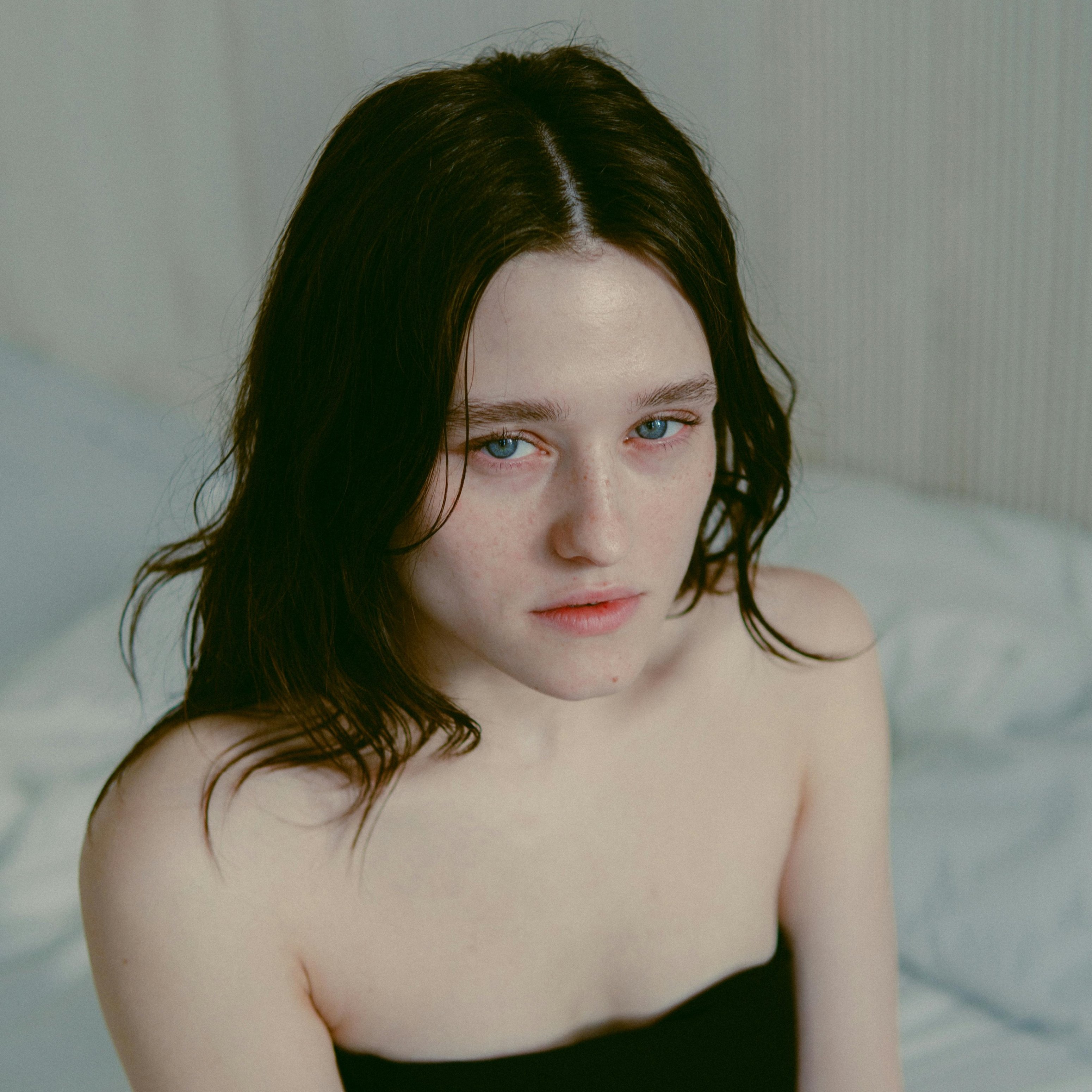 Porträt einer Frau mit blauen Augen und natürlicher Haut, die auf einem Bett sitzt, umgeben von einem hellen, minimalistischen Raum, passend zur Thematik der Hautpflege und Lösungen für geschwollene Augenlider.